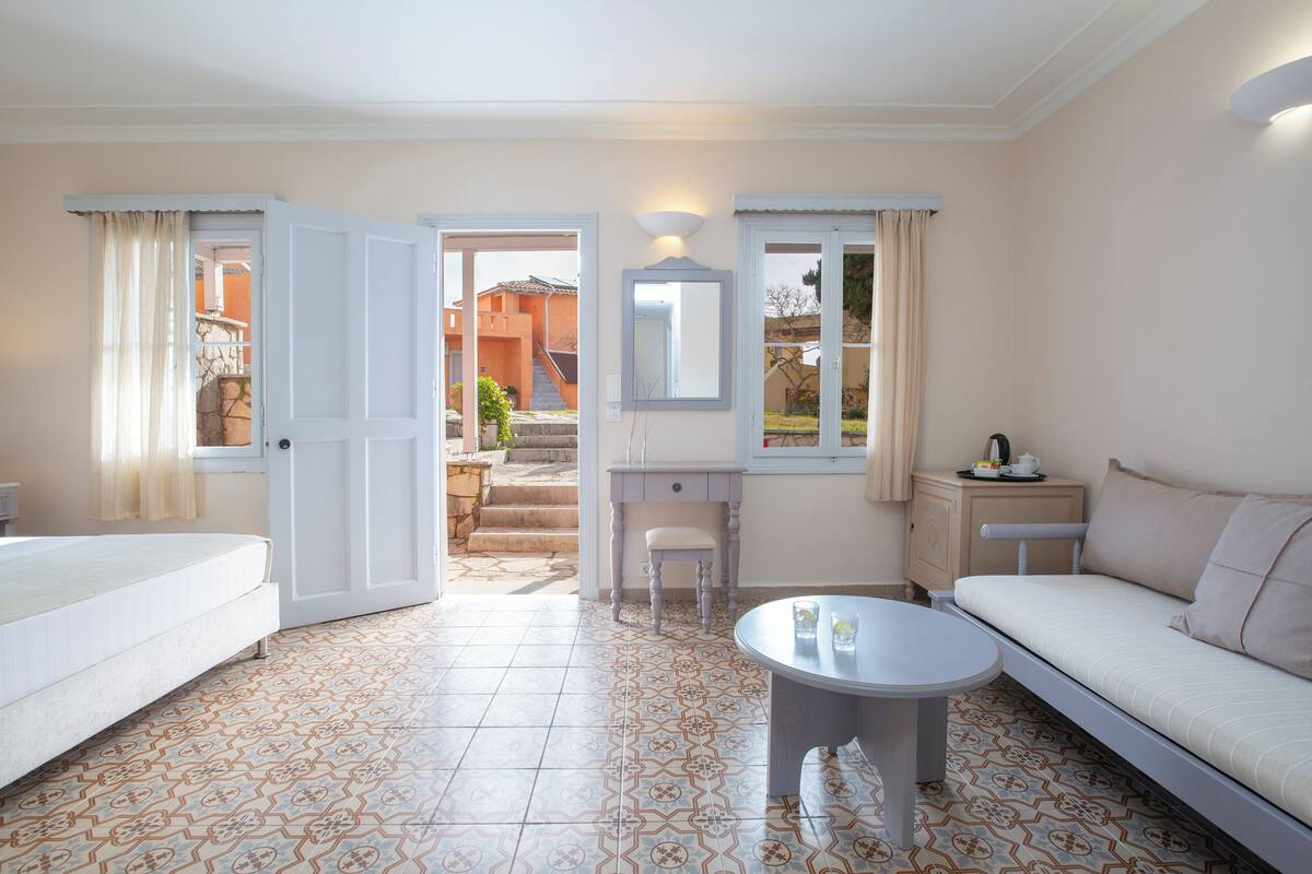 Quarto de hotel com uma cama, sofá, escrivaninha e cadeira. Há uma janela com cortinas, um espelho e uma pequena mesa.