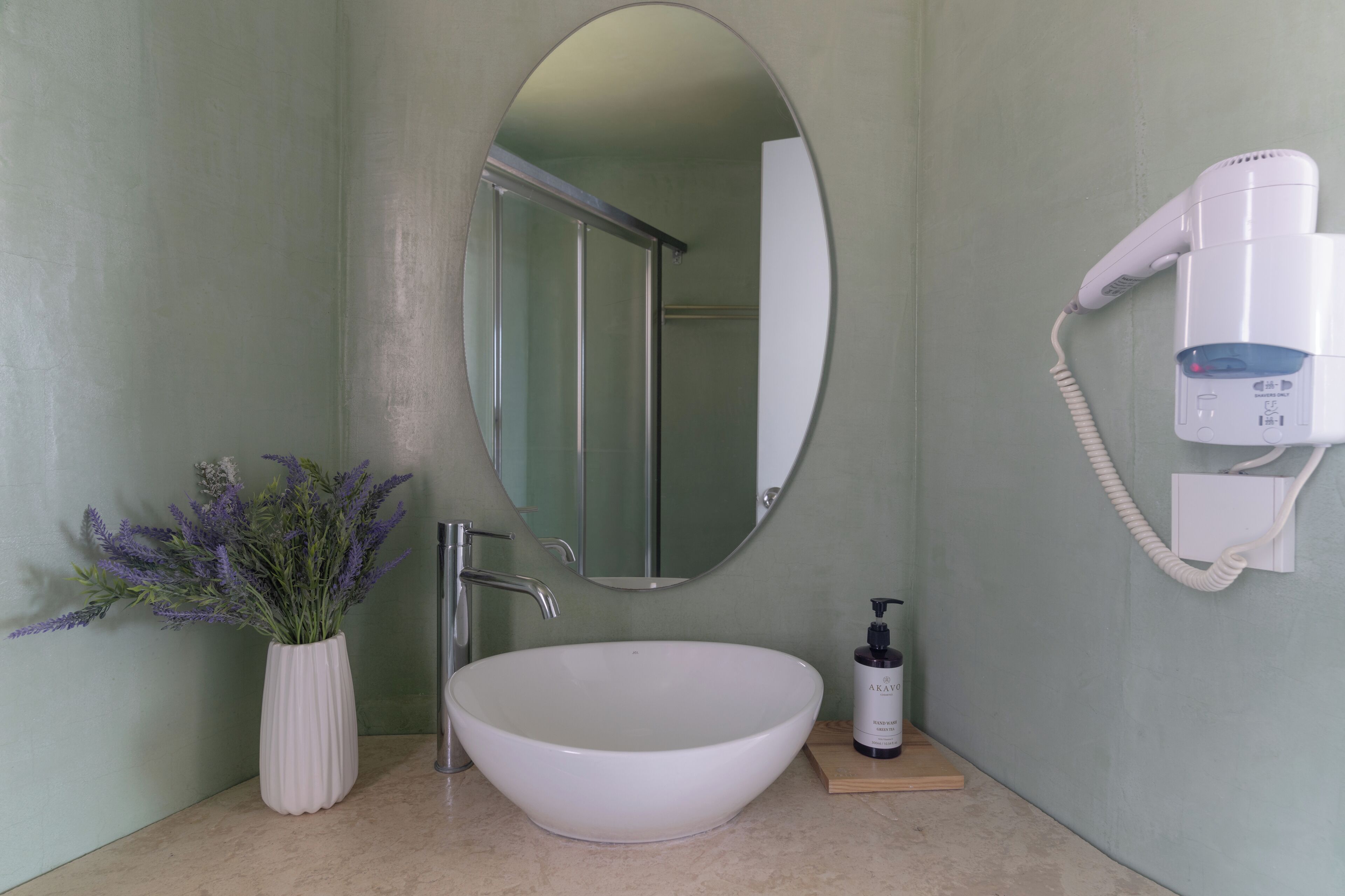 Une salle de bain avec un lavabo blanc, un miroir, un sèche-cheveux fixé au mur et un vase rempli de fleurs de lavande posé sur le comptoir.