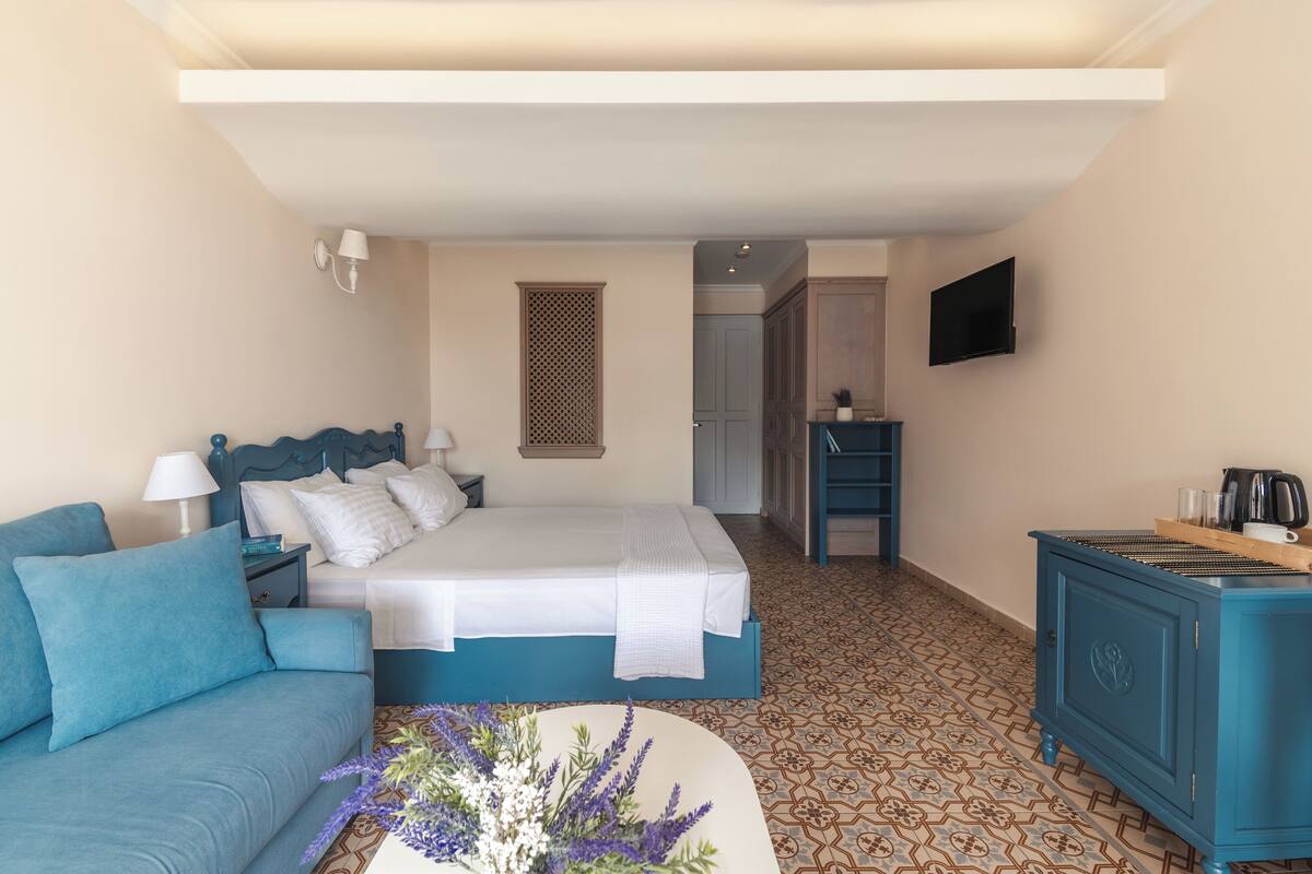 Quarto com cama, um sofá azul, mesa de cabeceira, cômoda e televisão.
