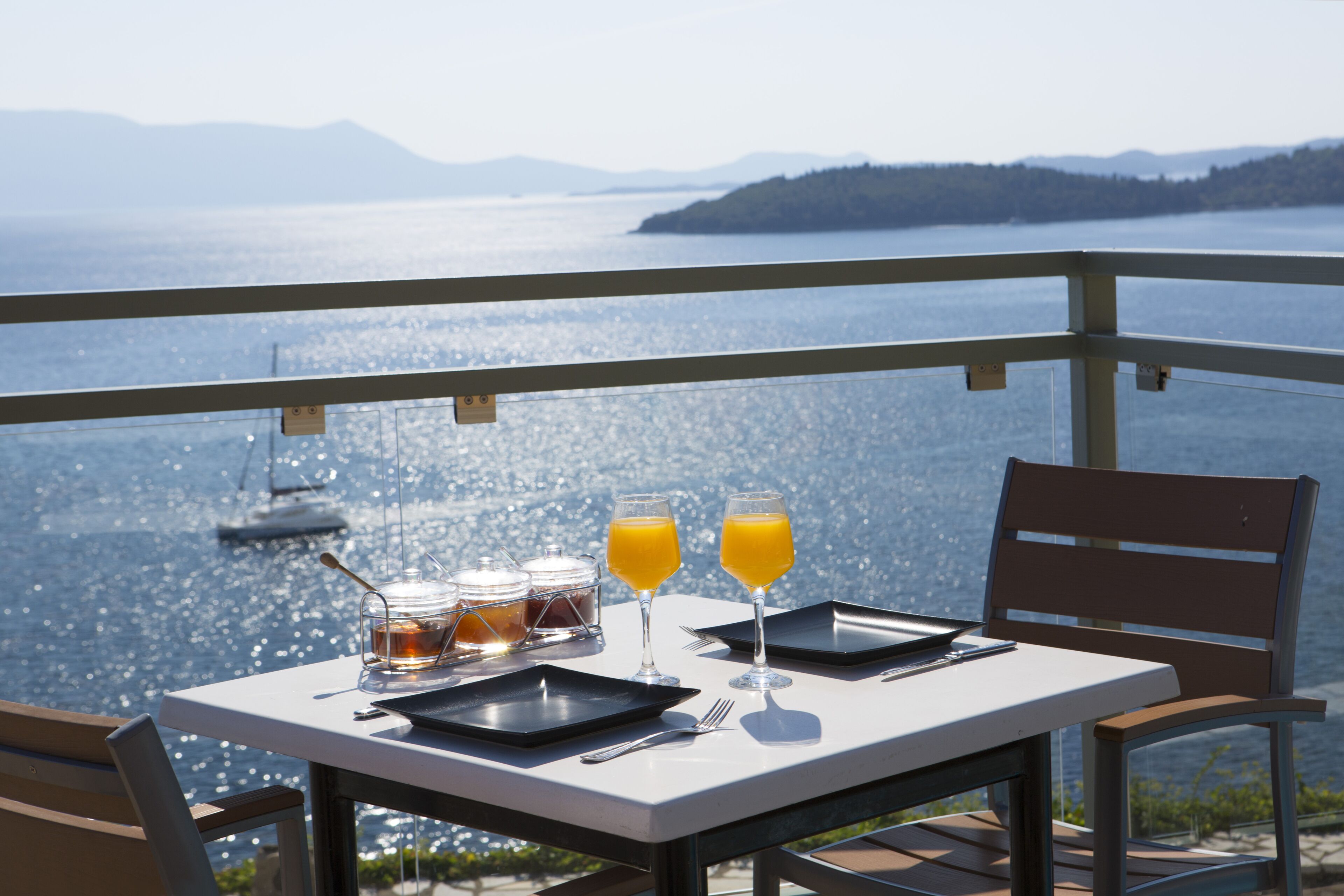 A table set with two glasses of orange juice, a jar of jam, and a bottle of water, overlooking a body of water with mountains in the distance.