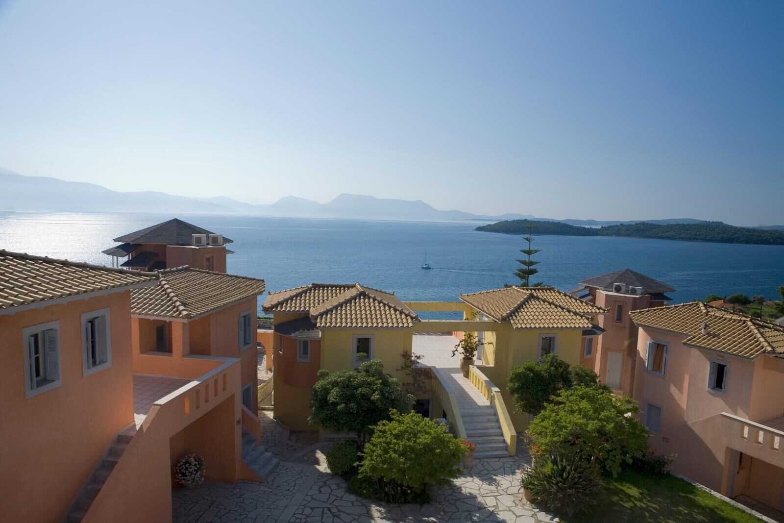 A coastal village with terracotta-roofed buildings, a stone pathway, and a view of the sea and mountains.