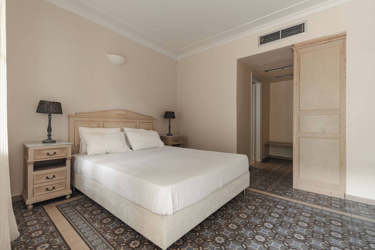 Quarto com cama, duas mesas de cabeceira com luminárias, guarda-roupa e piso com azulejos estampados.