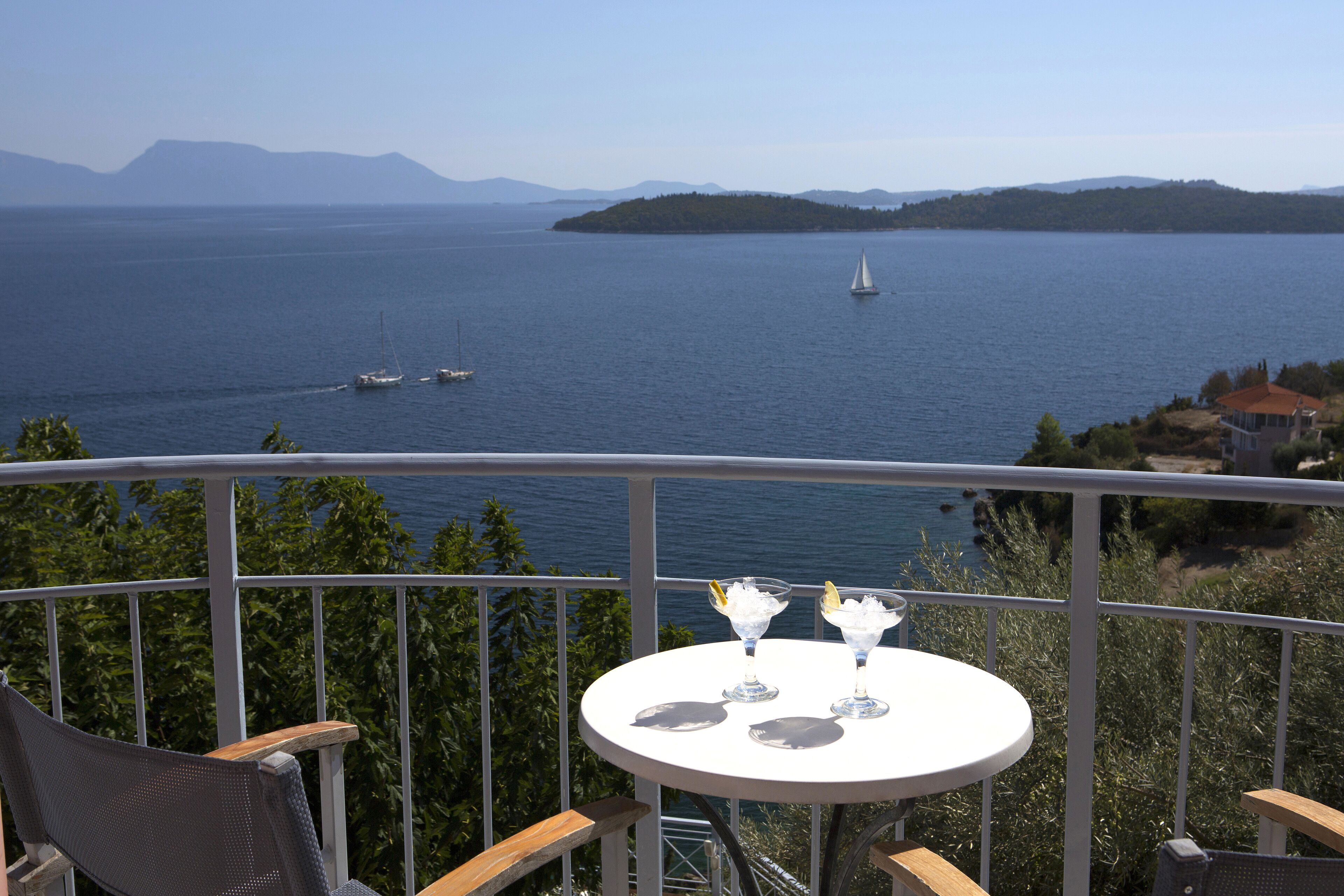 A balcony with a table set for two, overlooking a calm sea with boats and distant islands.