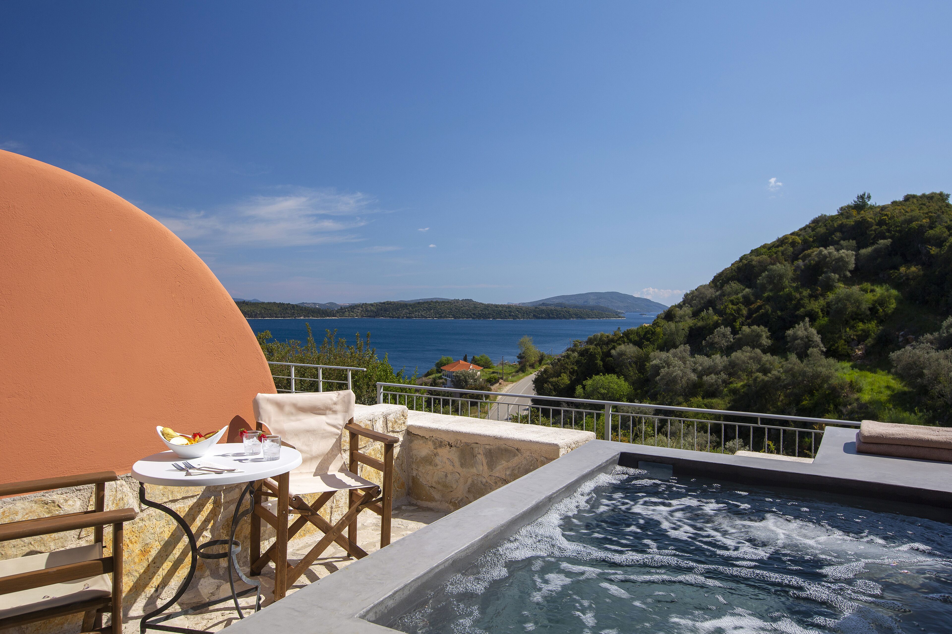A terrace with a hot tub, dining table, and chairs overlooking a scenic view of the sea and hills.