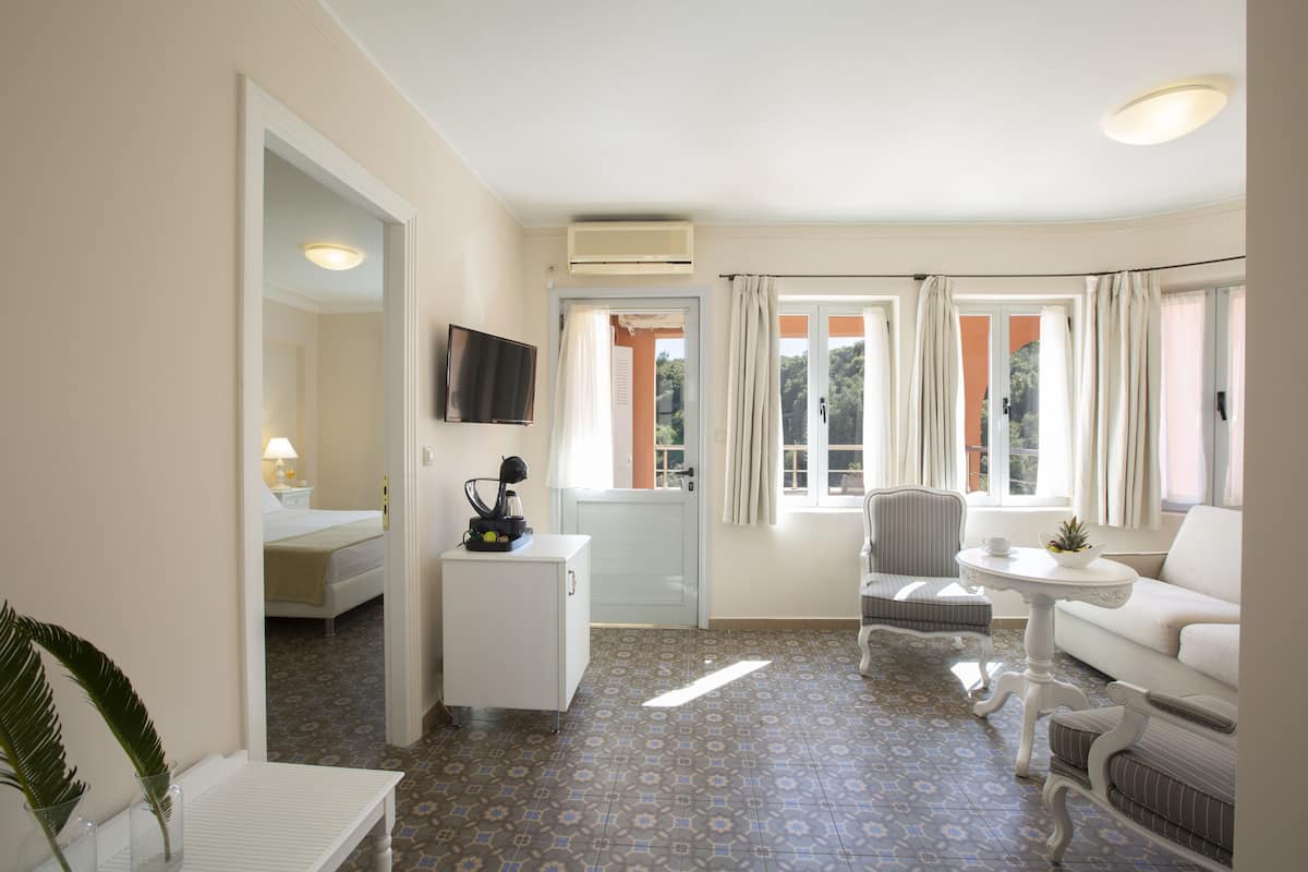 Quarto de hotel com cama, pequena escrivaninha, televisão, sofá, poltrona, mesa e janela com cortinas.