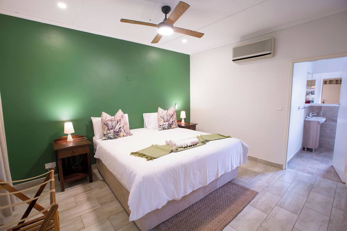 A neatly arranged bedroom with a green wall, a ceiling fan, a bedside table with a lamp, and a bathroom visible through an open door.