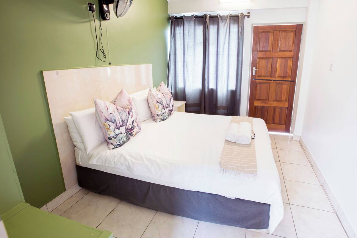 A neatly made bed with a white comforter and pillows, a wooden headboard, and a dark skirted bed base. There is a wooden door and a window with curtains in the background.