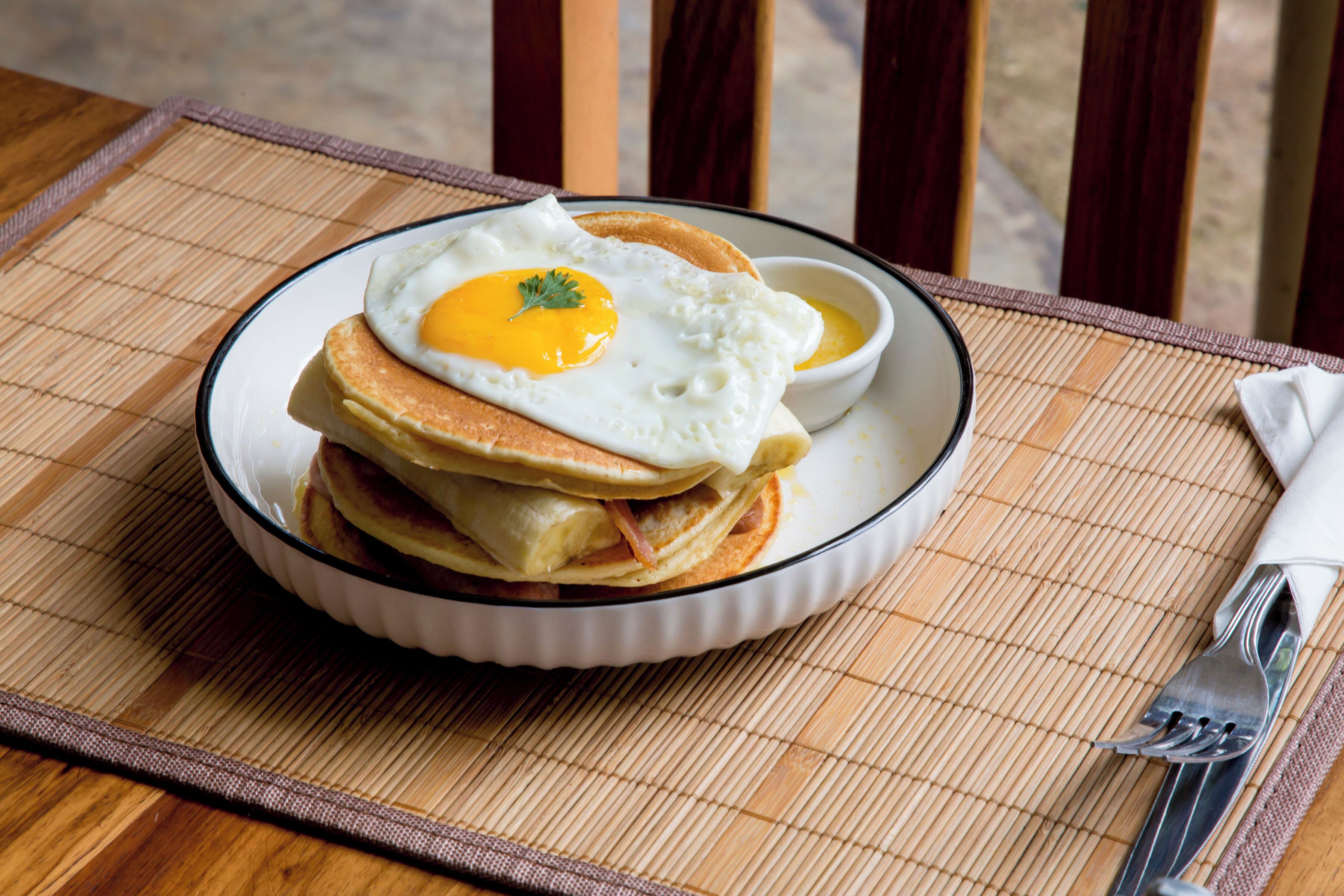 Un plat de crepes amb un ou de sol a sobre, presentat sobre un plat blanc amb vora negra, posat sobre un estores de bambú.