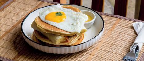 A plate of pancakes with a fried egg on top, served on a white plate with a black rim, placed on a bamboo mat.
