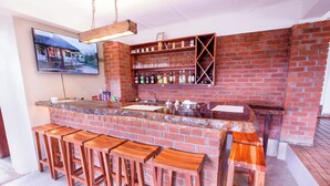 A bar area with a brick wall, wooden stools, a counter, and a shelf with bottles and glasses.