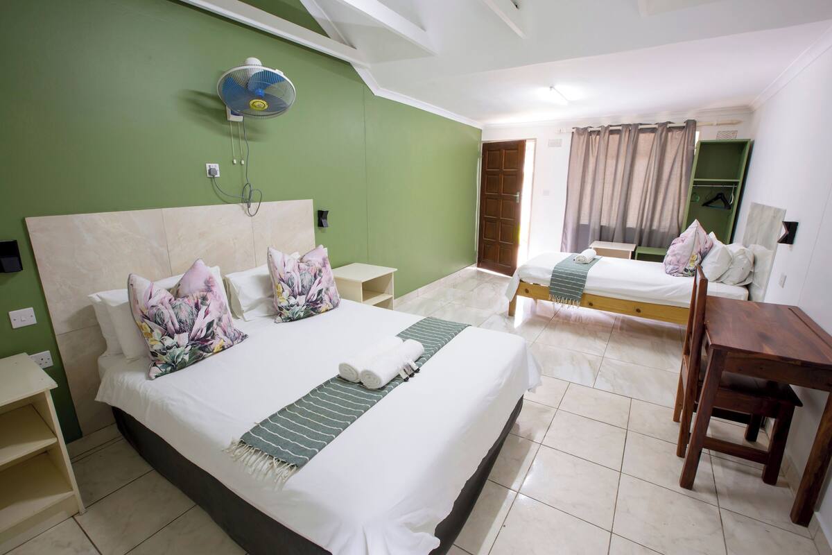 A hotel room with two beds, a wooden table, and a fan.