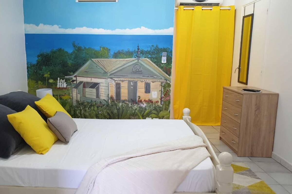 Un dormitorio con cama, almohadas, un aparador y un mural de una casa en la pared.