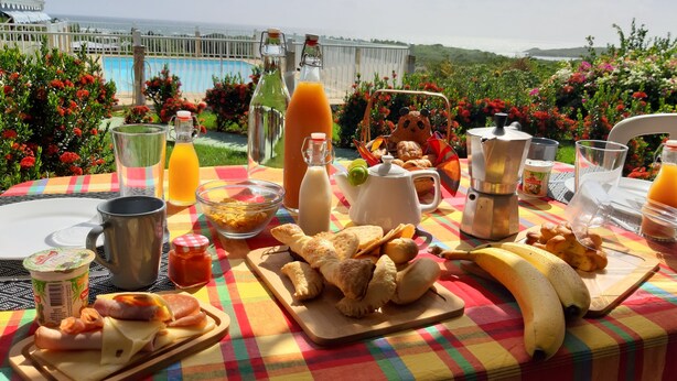 Una mesa de desayuno al aire libre con vista al mar, que incluye croissants, plátanos y diversas bebidas.