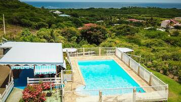 Una piscina rodeada por una valla blanca, con vista al océano y abundante vegetación al fondo.