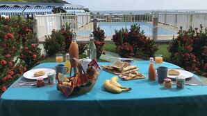 Montaje de comedor al aire libre con una mesa dispuesta para el desayuno, que incluye pan, frutas y bebidas, con vista a una piscina y un paisaje pintoresco.