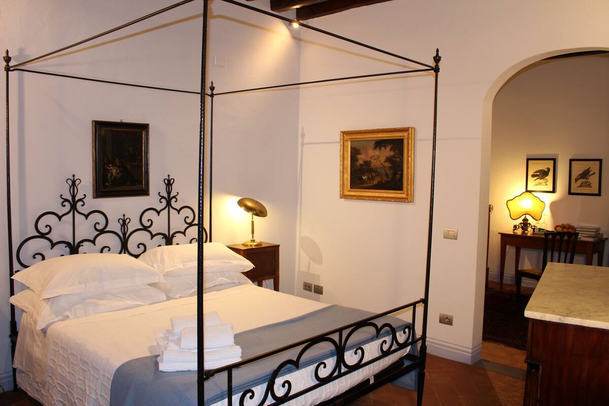 Una cama con dosel adornada con una colcha y almohadas blancas, una mesita de noche con lámpara, y cuadros enmarcados en las paredes.