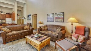 Ein geräumiges Wohnzimmer mit zwei braunen Ledersofas, einem Holztisch und einem grauen Sessel. An den Wänden sind zwei Lampen angebracht und über den Sofas hängen gerahmte Bilder.