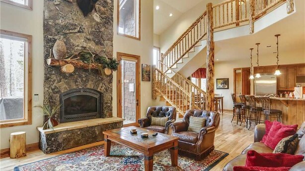 A spacious living room with a stone fireplace, leather sofas, a wooden staircase, and a kitchen area.