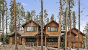 A two-story wooden cabin with a porch, surrounded by tall pine trees.
