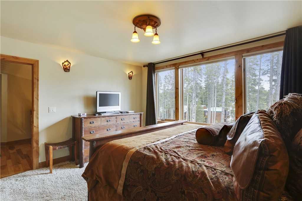 A bedroom with a large bed, a dresser, a television, and a view of trees through large windows.