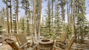 A wooden fire pit surrounded by four rustic chairs in a forest setting.