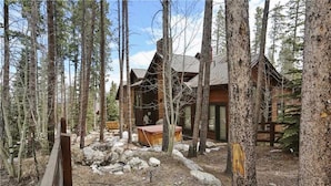 A cabin surrounded by tall trees, with a wooden deck and a hot tub in the foreground.