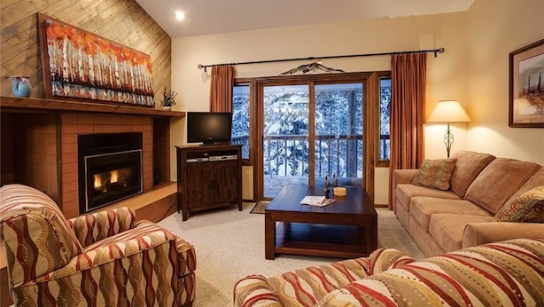 A living room with a fireplace, a television, a sofa, a coffee table, and a balcony with a view of trees. - Winterwood 3 Town House (Steamboat Springs)