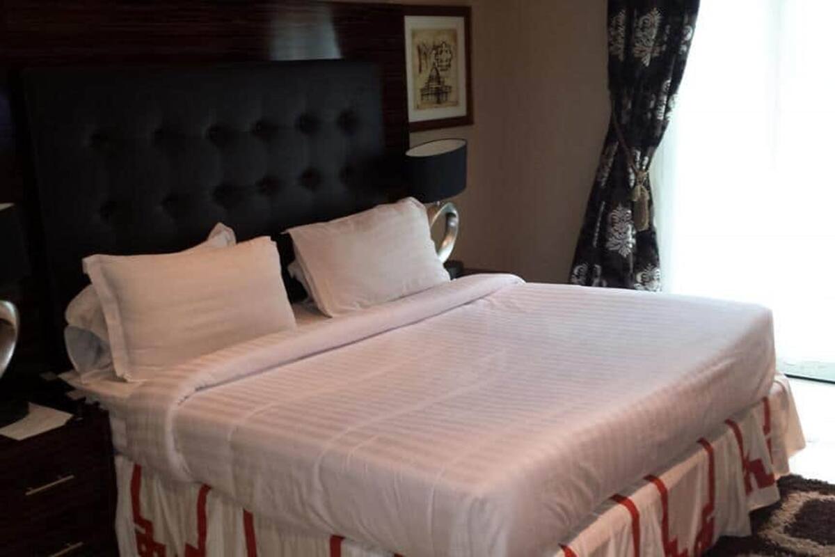 A neatly made bed with white linens and red accents, a dark upholstered headboard, a bedside table with a lamp, and a framed picture on the wall.