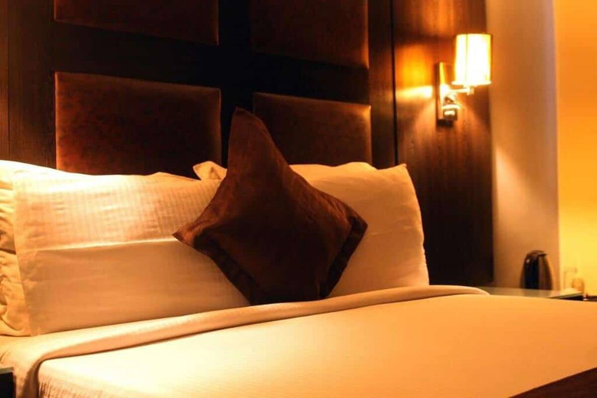 A hotel room with a bed, pillows, a nightstand, and a wall-mounted lamp.