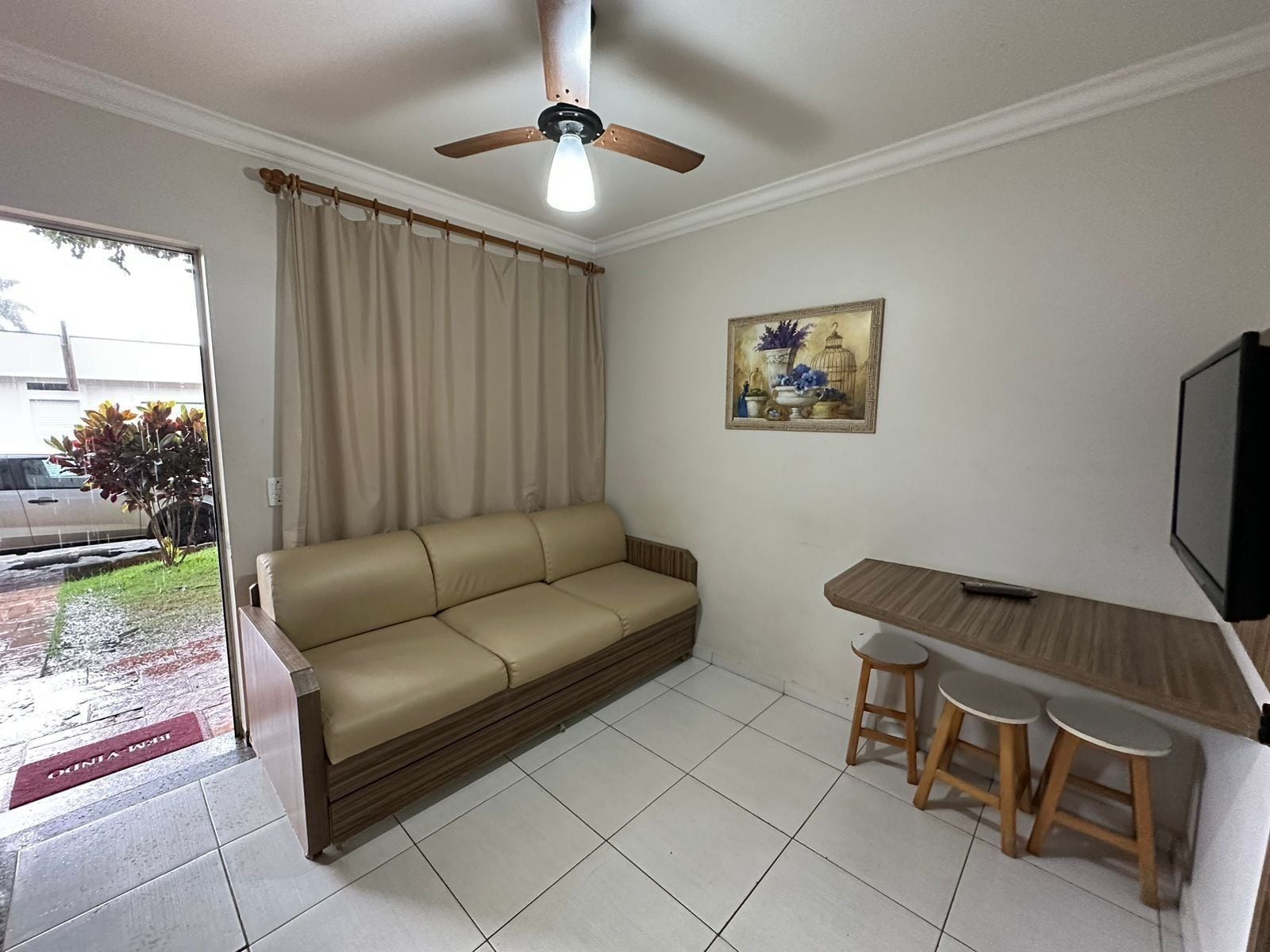 A living room with a sofa, a ceiling fan, a painting, a TV, and a small table with stools.