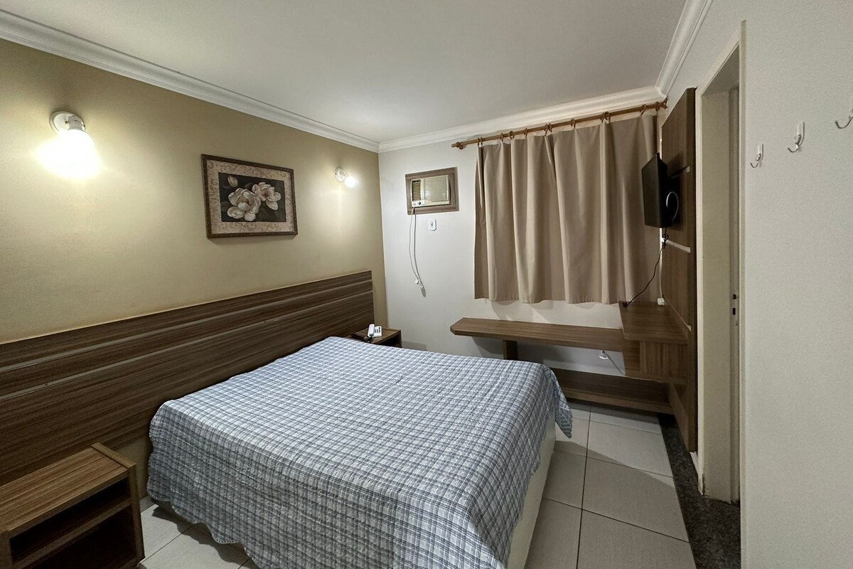 Quarto de hotel com cama, mesa de cabeceira, televisão na parede e janela com cortinas.