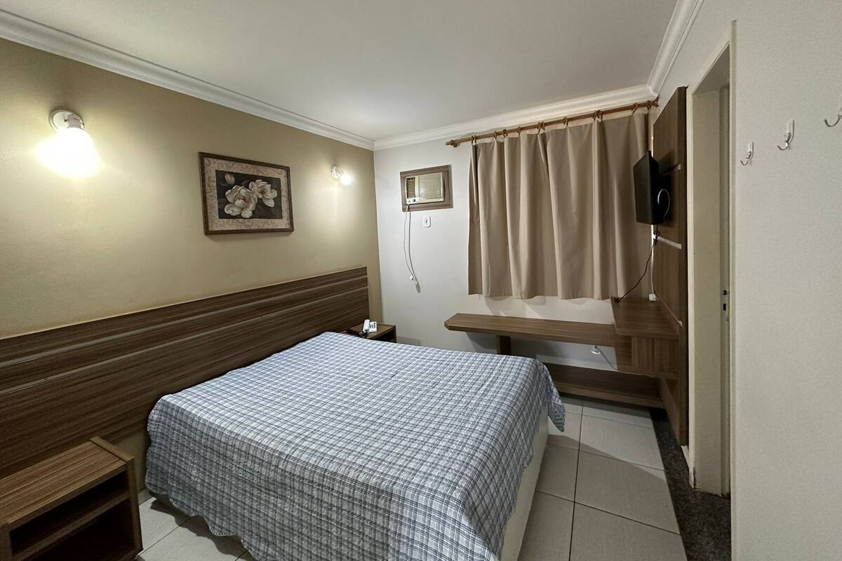 Quarto de hotel com cama, mesa de cabeceira, televisão na parede e janela com cortinas.