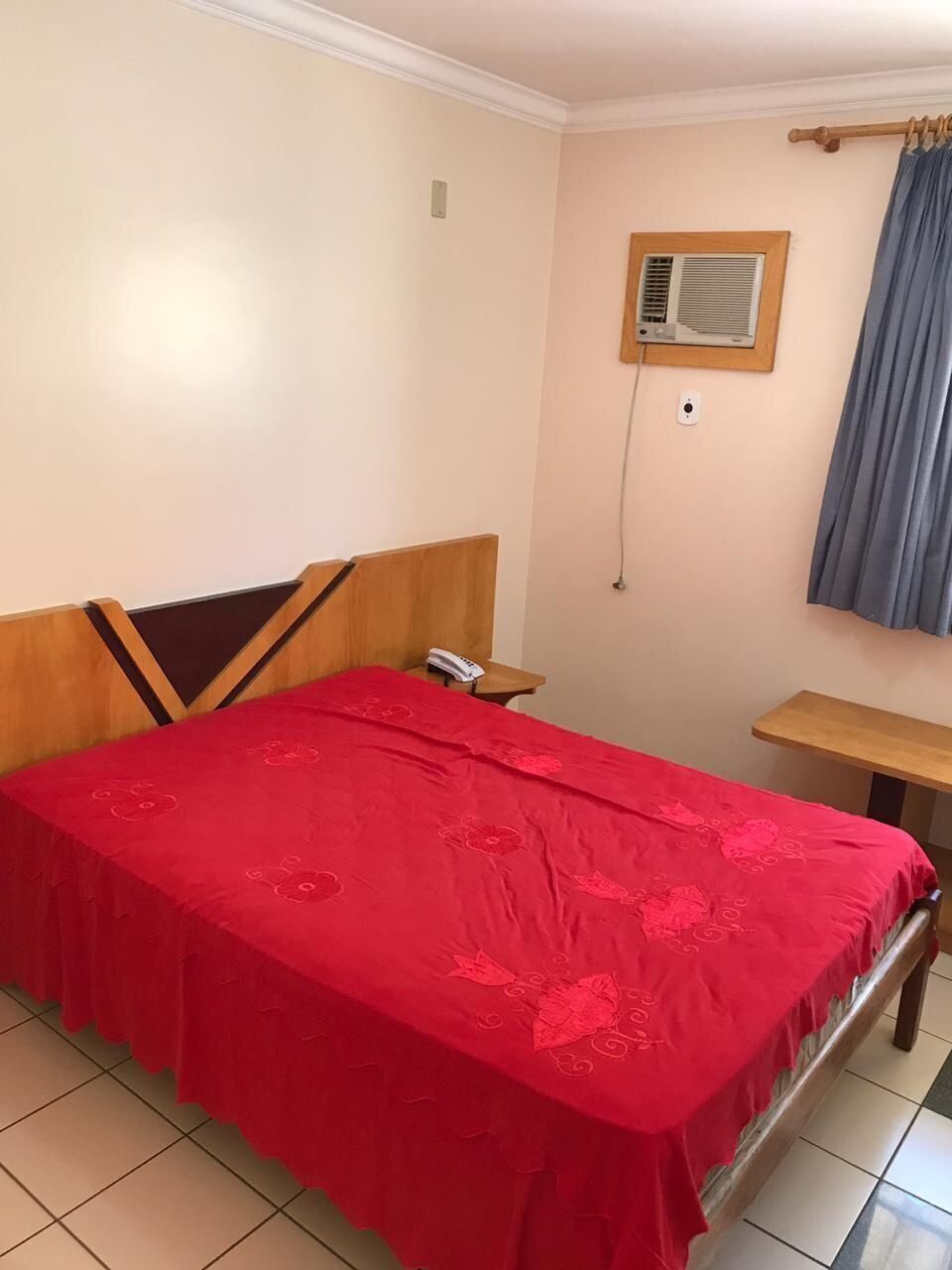Een slaapkamer met een bed, nachtkastje, telefoon, wandairconditioner en een raam met gordijnen.
