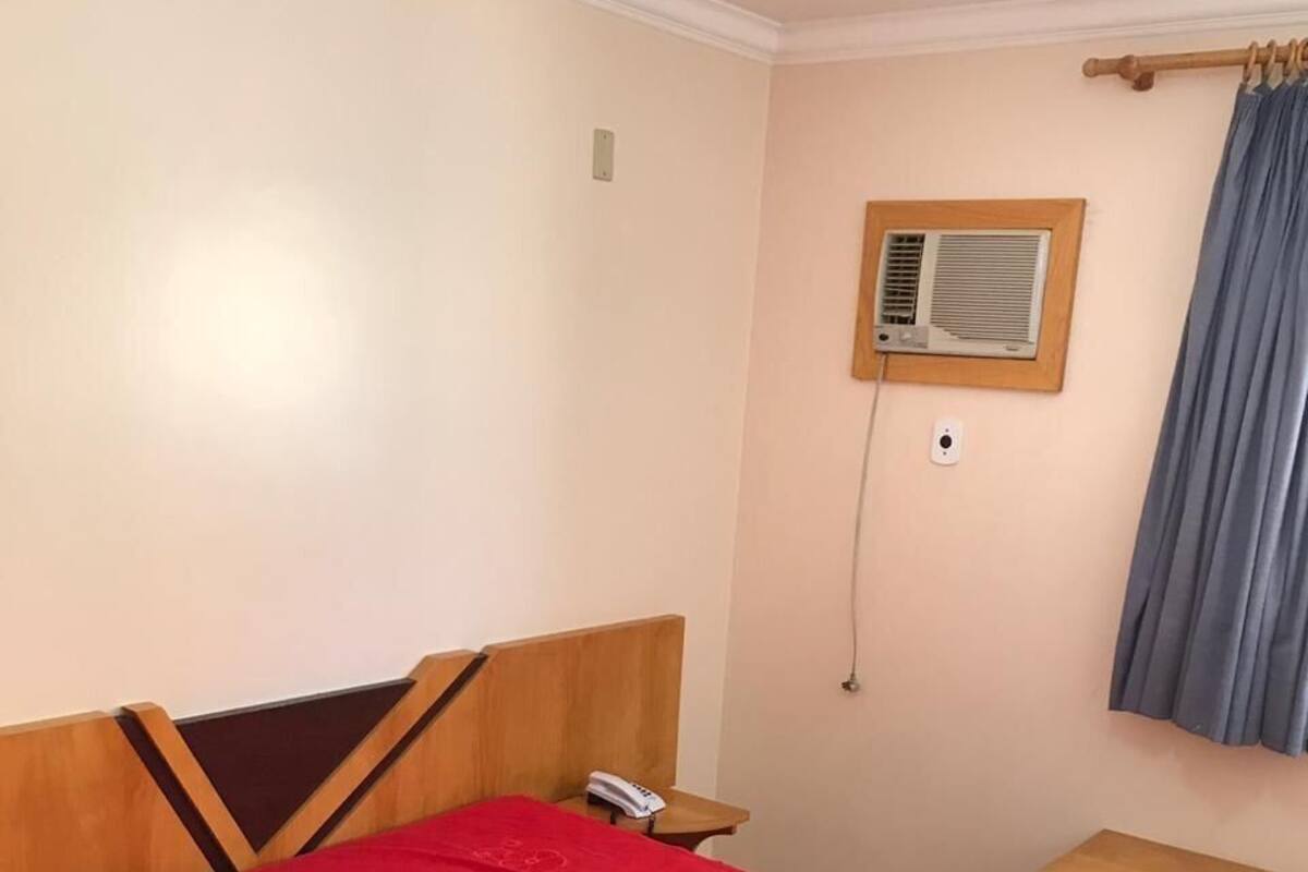 Quarto com cama, mesa de cabeceira, telefone, ar-condicionado na parede e janela com cortinas.