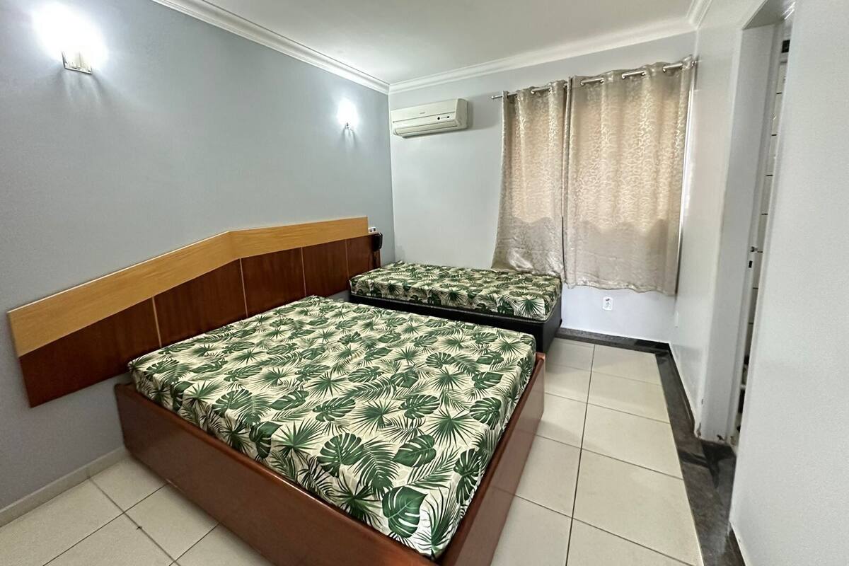 Quarto com duas camas, janela com cortinas, ar condicionado e luminária fixada na parede.