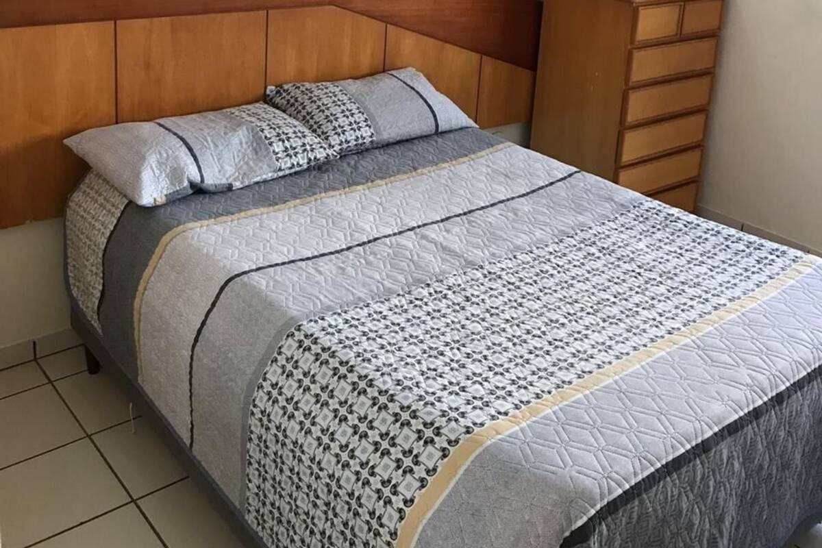 Quarto com cama, cabeceira de madeira, cômoda e ar-condicionado fixado na parede.