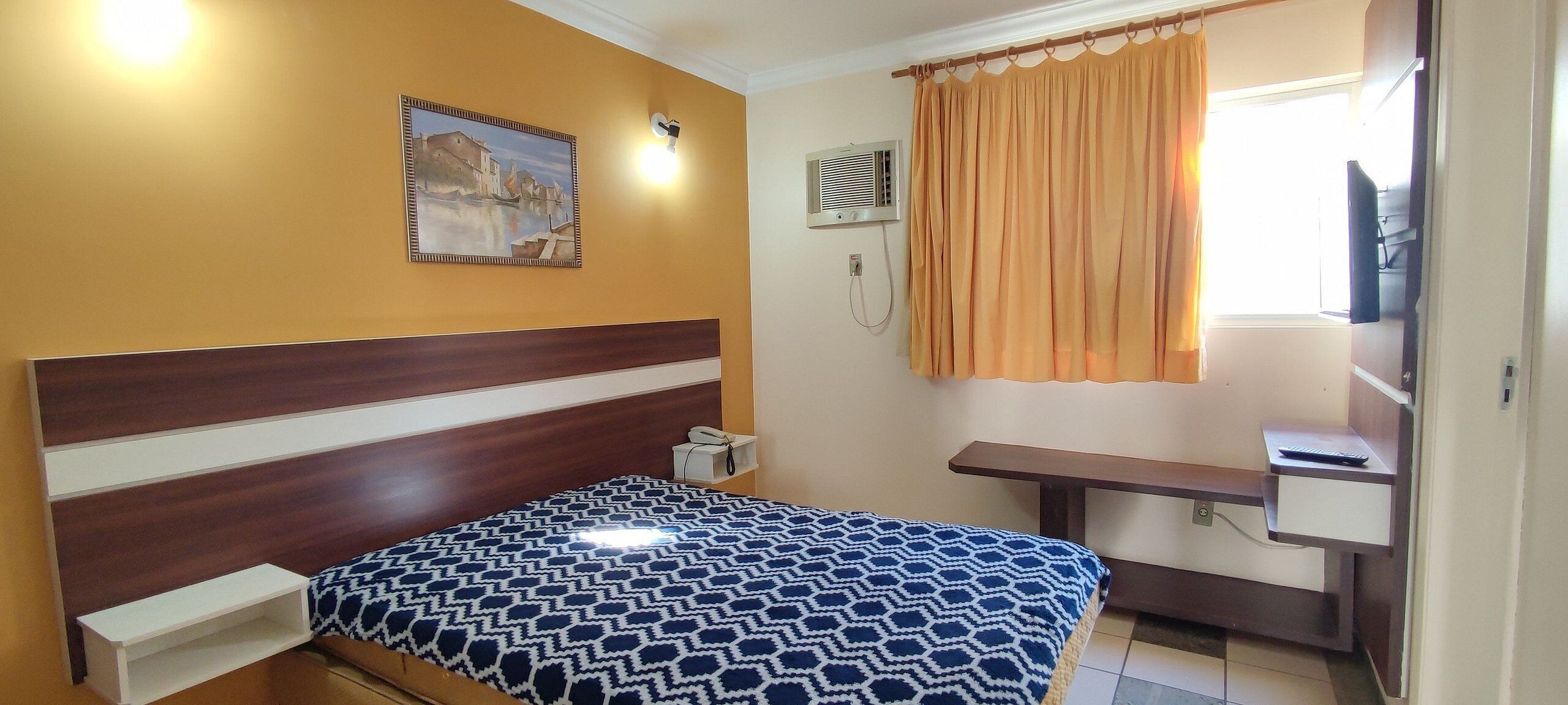 Hotelkamer met een bed, bureau, stoel, raam met gordijnen, een luchtverkoeler aan de muur en een schilderij.