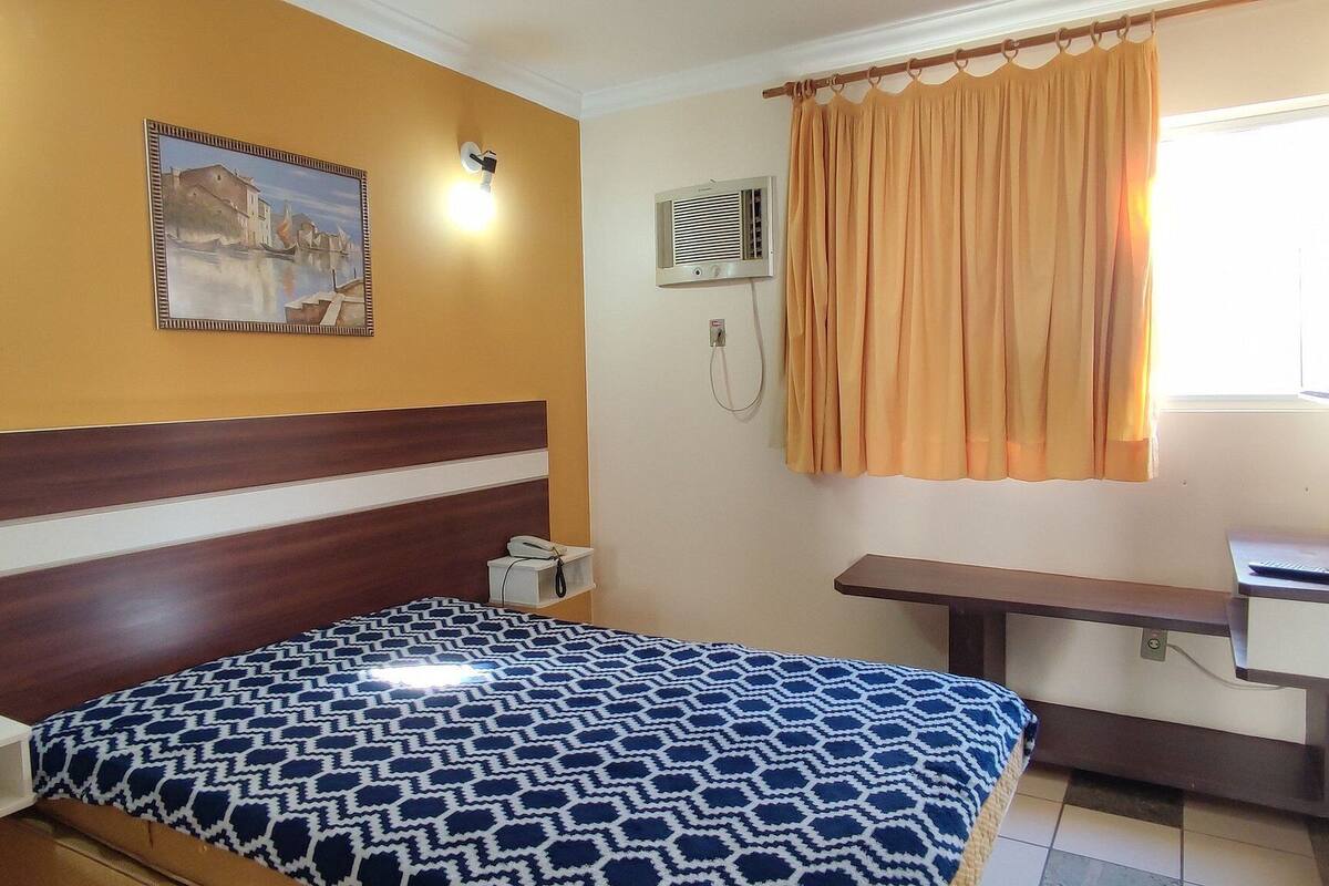 Quarto de hotel com cama, escrivaninha, cadeira, janela com cortinas, ar condicionado fixado na parede e um quadro.