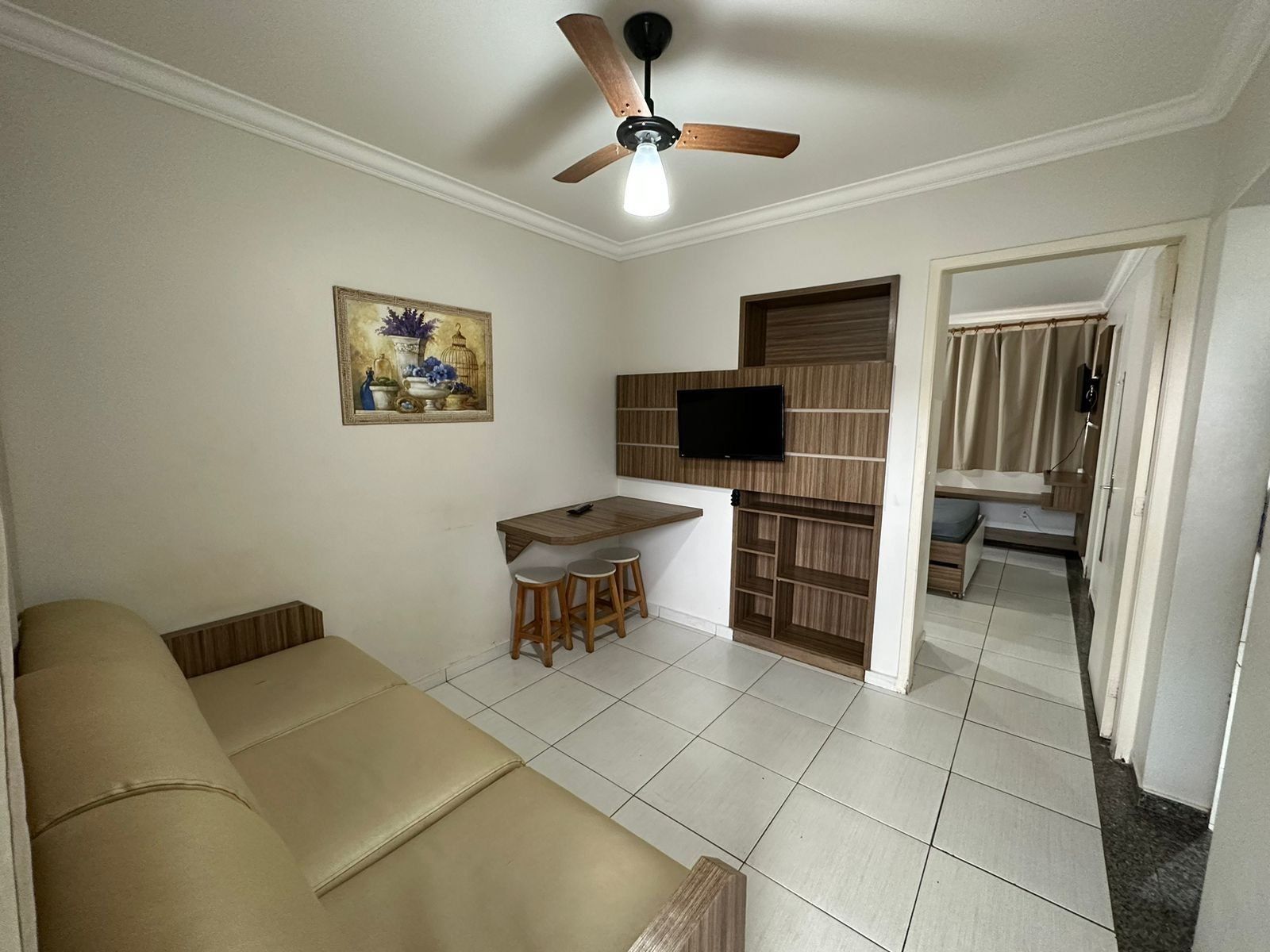 Una sala de estar con un sofá, una zona de comedor con taburetes, un televisor montado en un panel de madera y un ventilador de techo.
