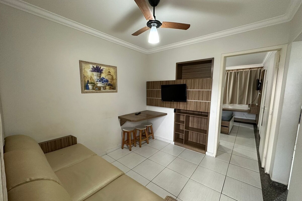Ein Wohnzimmer mit einer Couch, einem Essbereich mit Hocker, einem an einer Holzvertäfelung montierten Fernseher und einem Deckenventilator.
