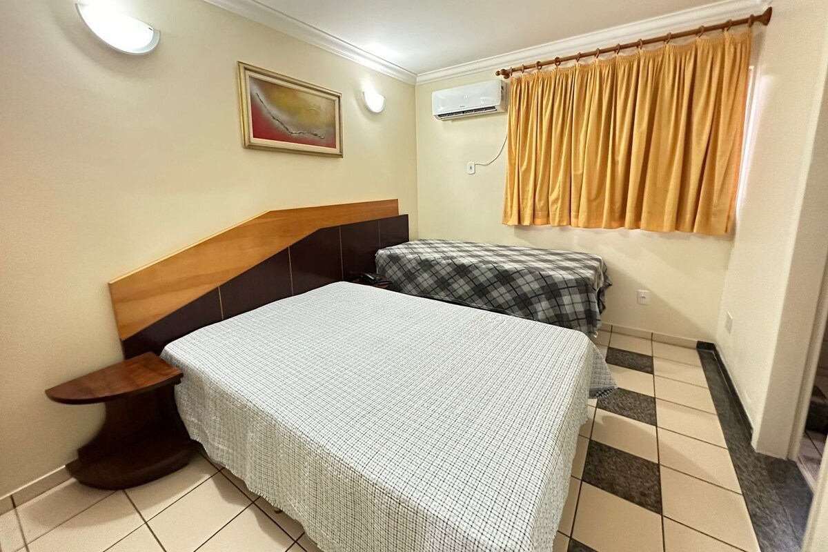 Quarto com cama, mesa de cabeceira, ar condicionado fixado na parede, janela com cortinas e um quadro na parede.