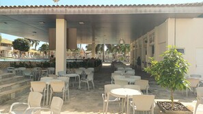 An outdoor cafe area with tables and chairs, a pool in the background, and a covered walkway. - DiRoma International Resort Via Caldas (Caldas Novas)