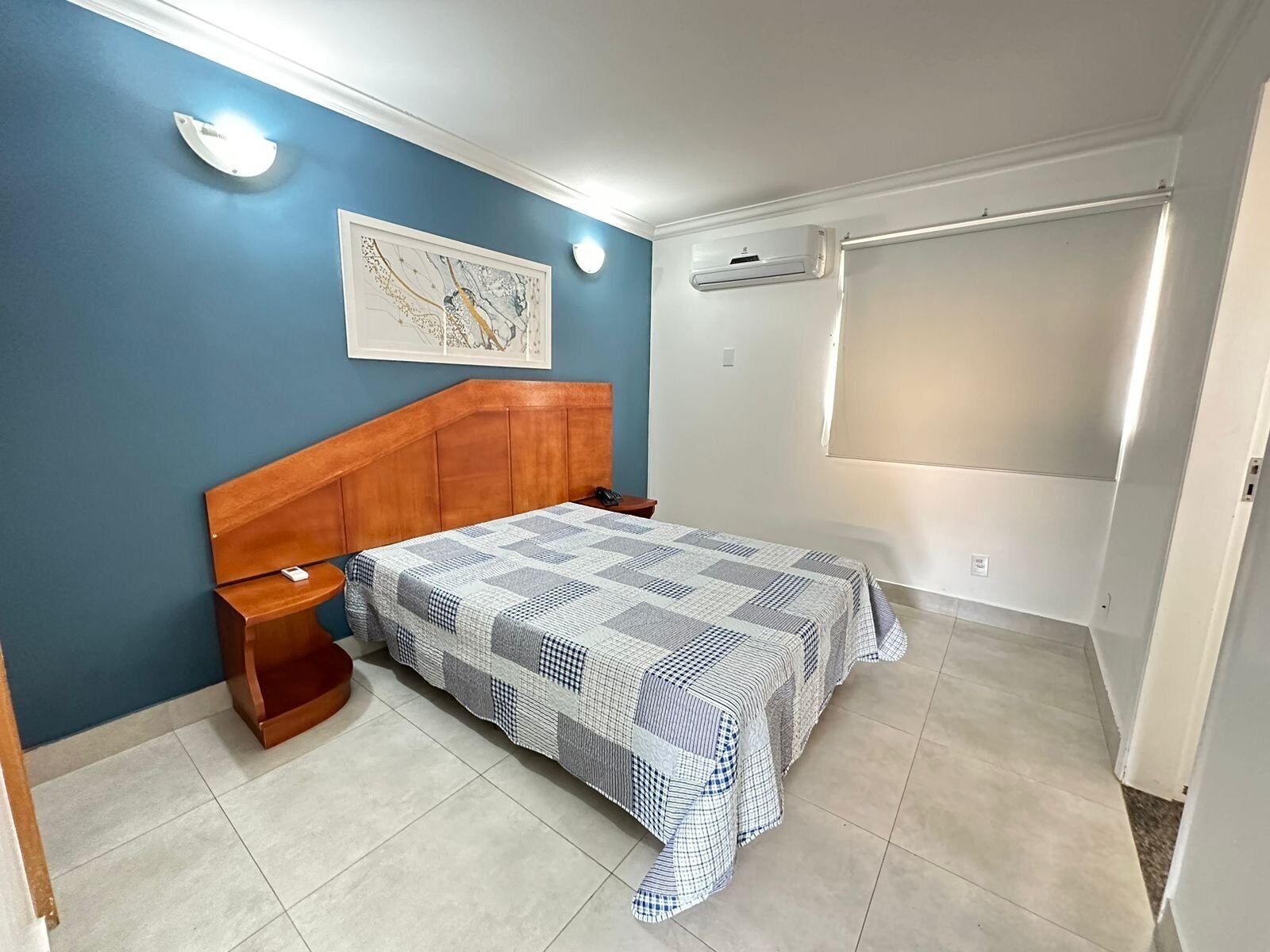 Een slaapkamer met een houten hoofdbord, een geruit beddegoed, een nachtkastje, een airconditioner en een lijst met een kunstwerk aan de muur.