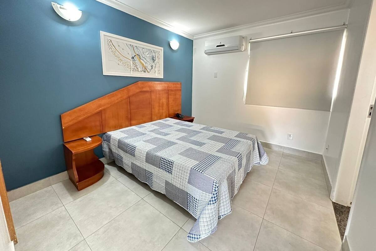 Quarto com cabeceira de madeira, colcha xadrez, mesa de cabeceira, ar condicionado e uma obra de arte emoldurada na parede.