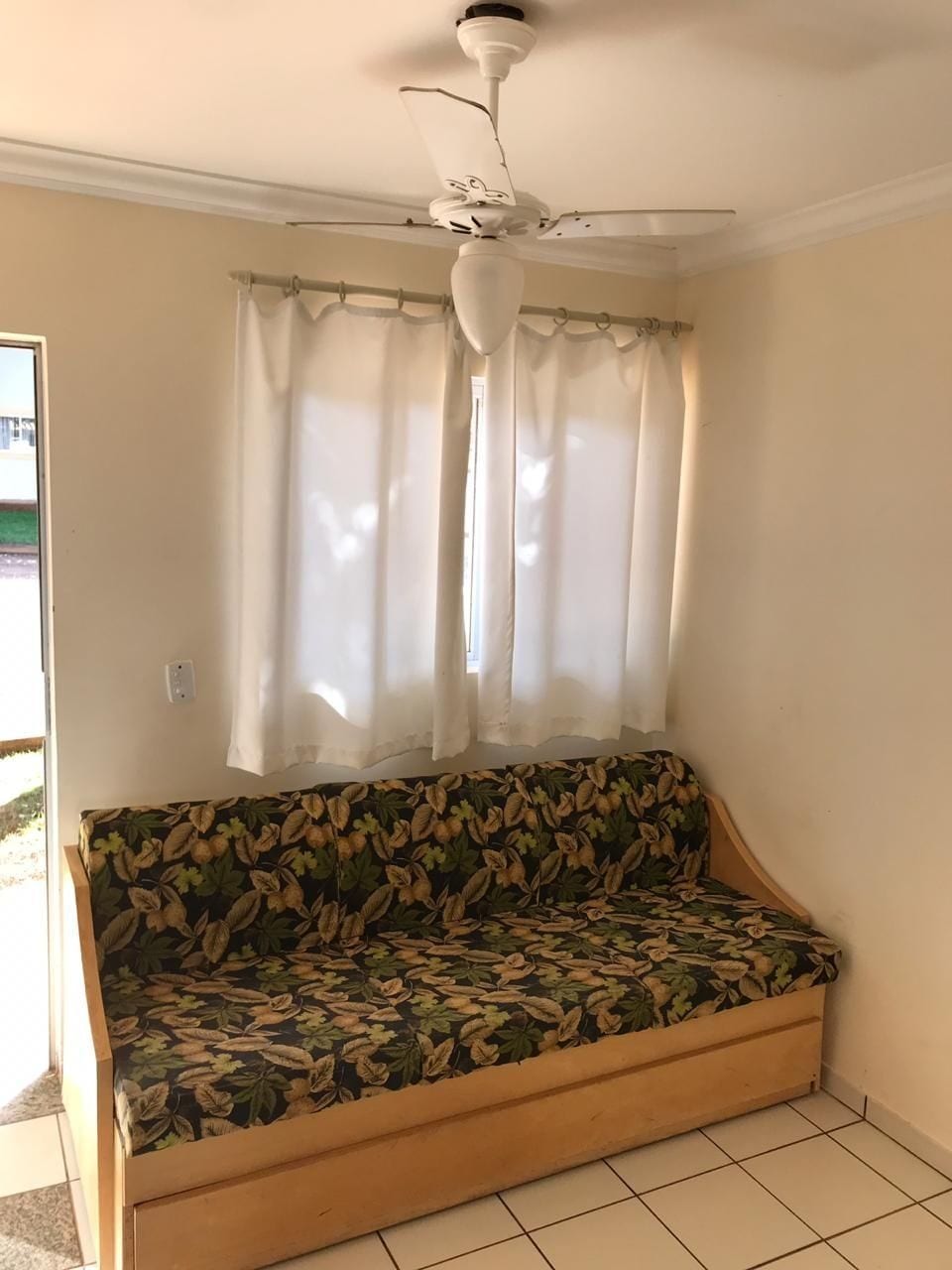 A room with a ceiling fan, a sofa with a floral pattern, and a window with sheer curtains.