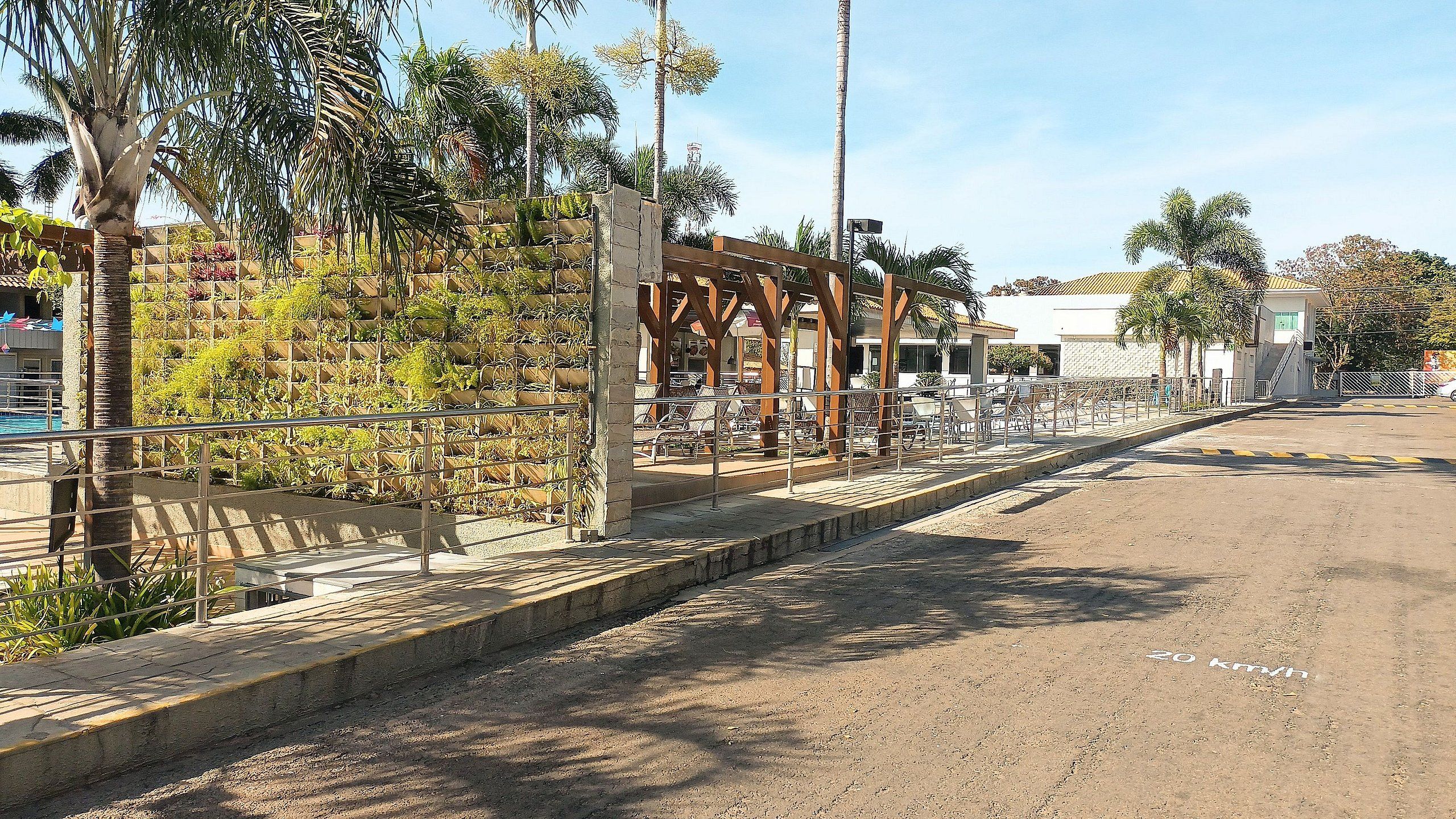 A paved walkway with a metal railing, palm trees, and a building with a white facade in the background.