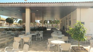 Ein Außenbereich mit Café, Tischen, Stühlen und einem Pool im Hintergrund sowie einem überdachten Gang.