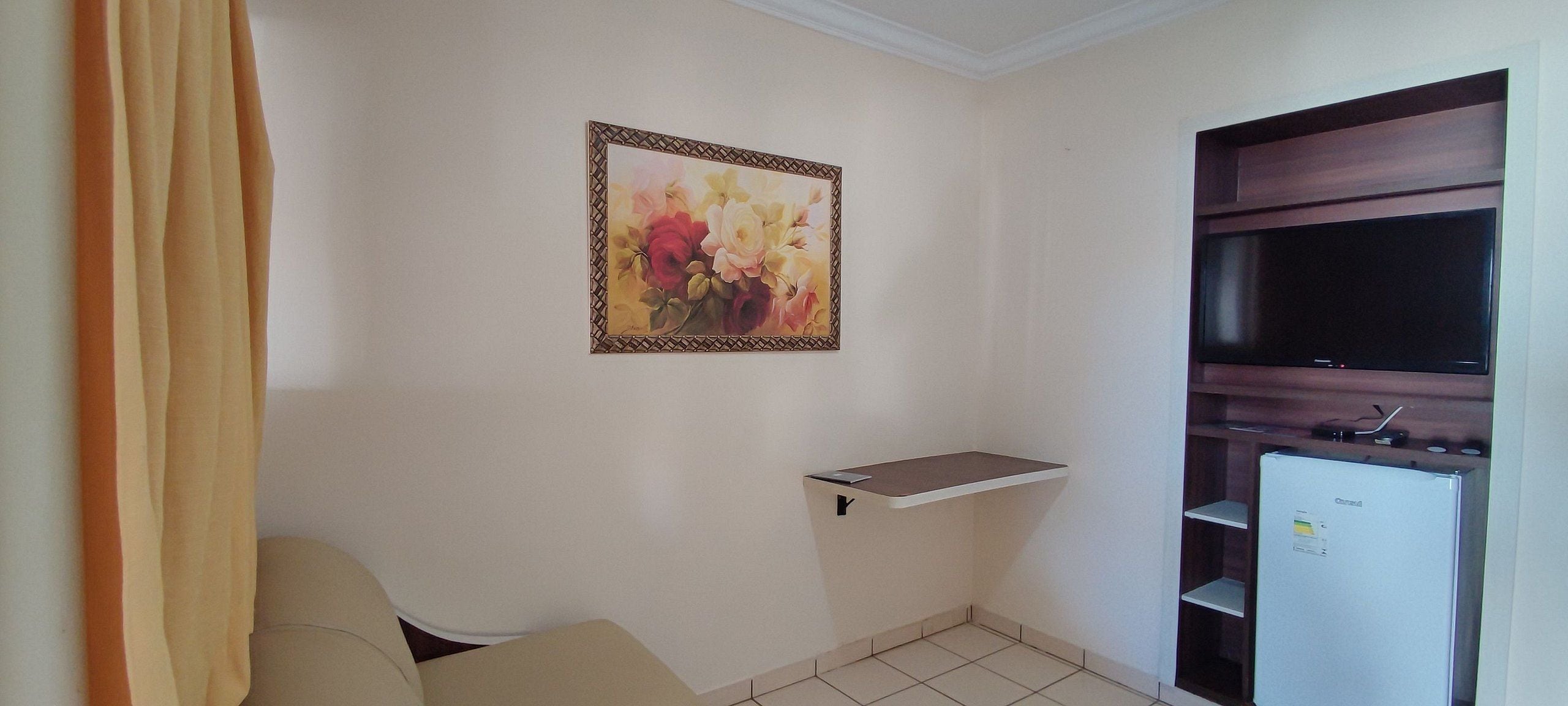 A room with a flat-screen TV, a small white refrigerator, a beige sofa, a wall-mounted shelf, and a painting of flowers.