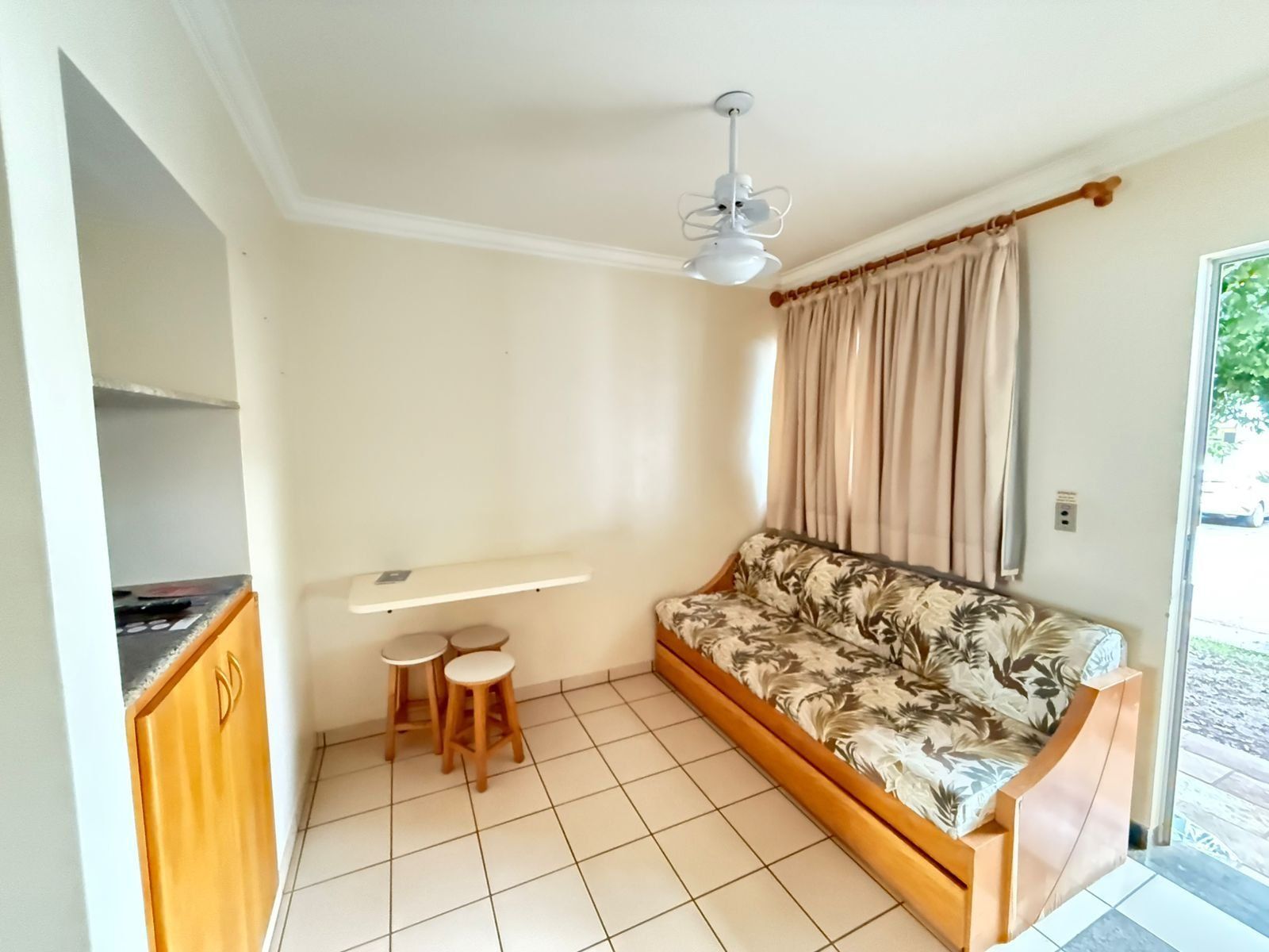 Quarto com sofá, ventilador de teto, pequena mesa com banquetas, janela com cortinas e porta que dá para o exterior.