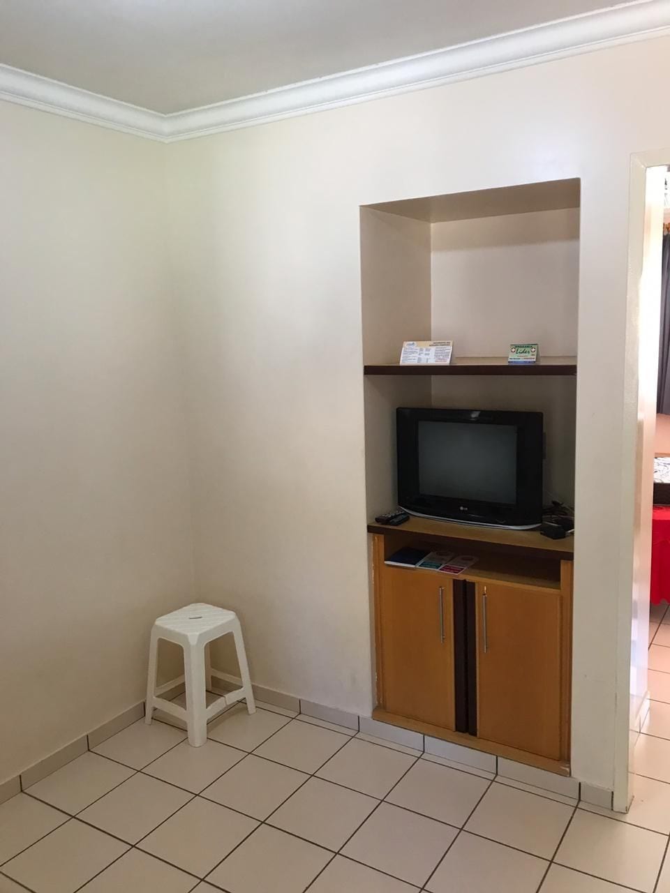 A room with a television on a wooden stand, a white stool, and a cabinet with a red table in the background.